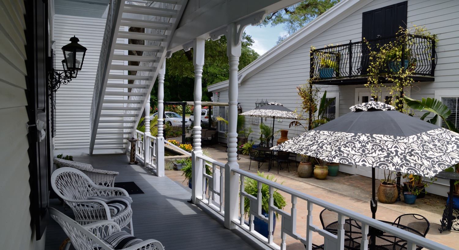 Covered porch, white posts, railing and chairs, painted wood floor, stairs to upper rooms, courtyard with patio tables with umbrellas