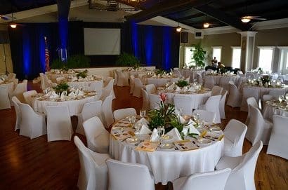 Ballroom set-up corporate style with white linens and chair covers including place settings and audio visual set-up.