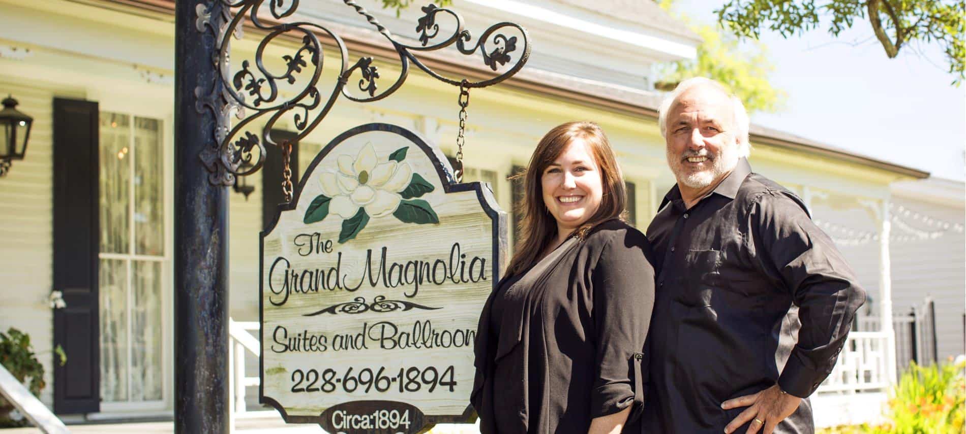 Owners, a man and woman, smiling outside standing next to the a hanging sign that says: The Grand Magnolia Suites and Ballroom 228-696-1894 Circa 1894