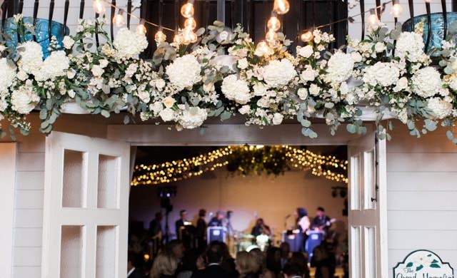 Double glass French doors swung open with large white flowers hanging overhead with view of wedding band and twinkle lights inside