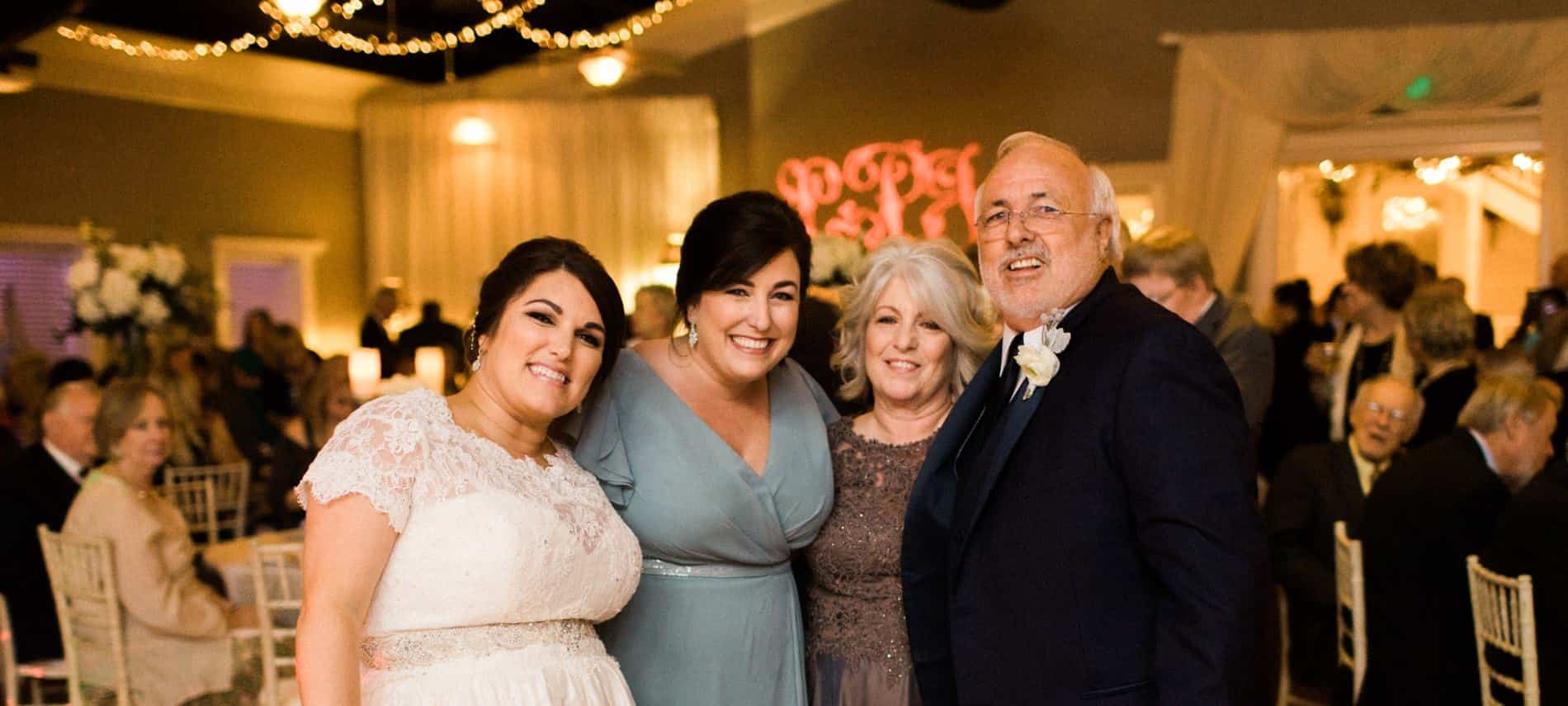 Bride and groom with two women all smiling amidst a crowd of people at tables and chairs with twinkle lights hanging above