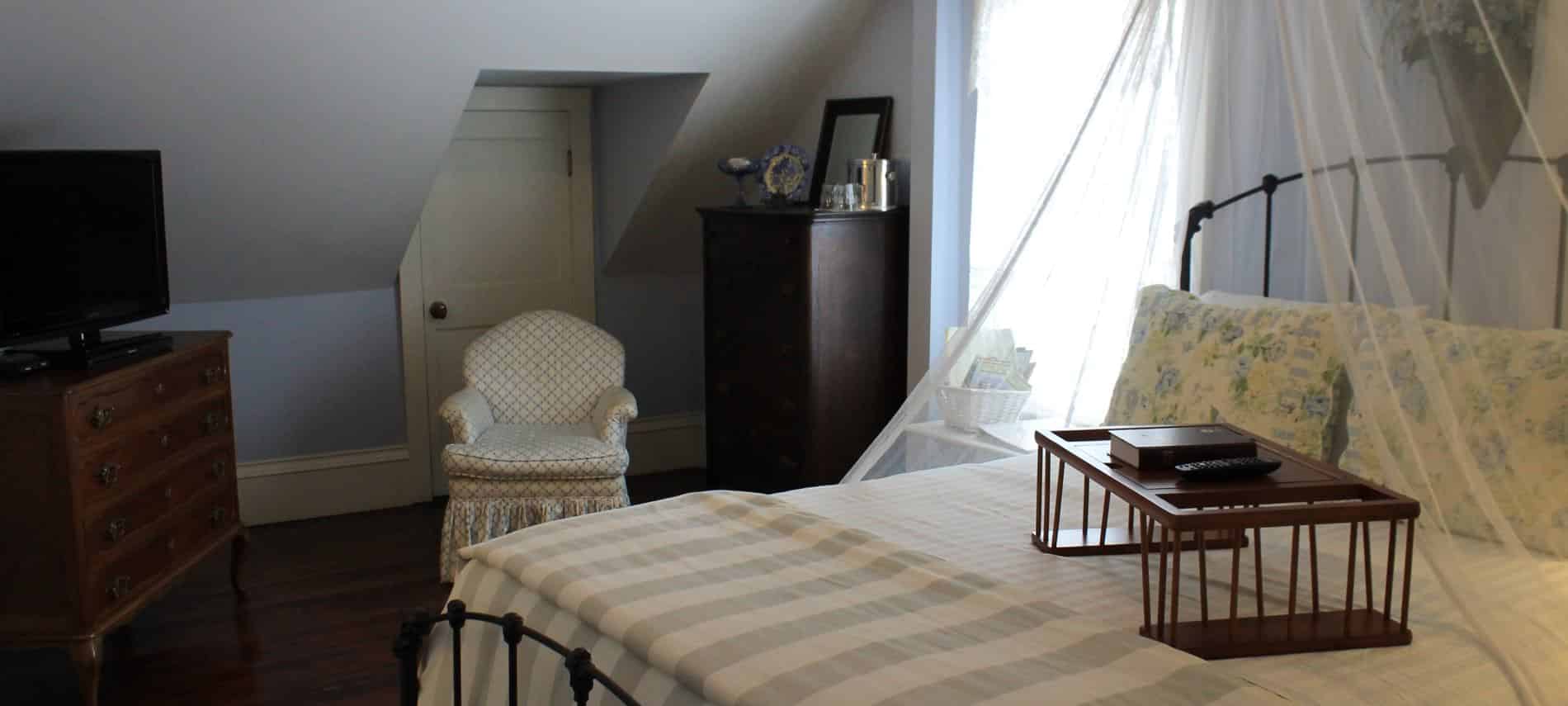 Cape Cod guest room, slanted ceiling, wood floor, window, metal bed with sheer white canopy, upholstered chair, dresser and TV