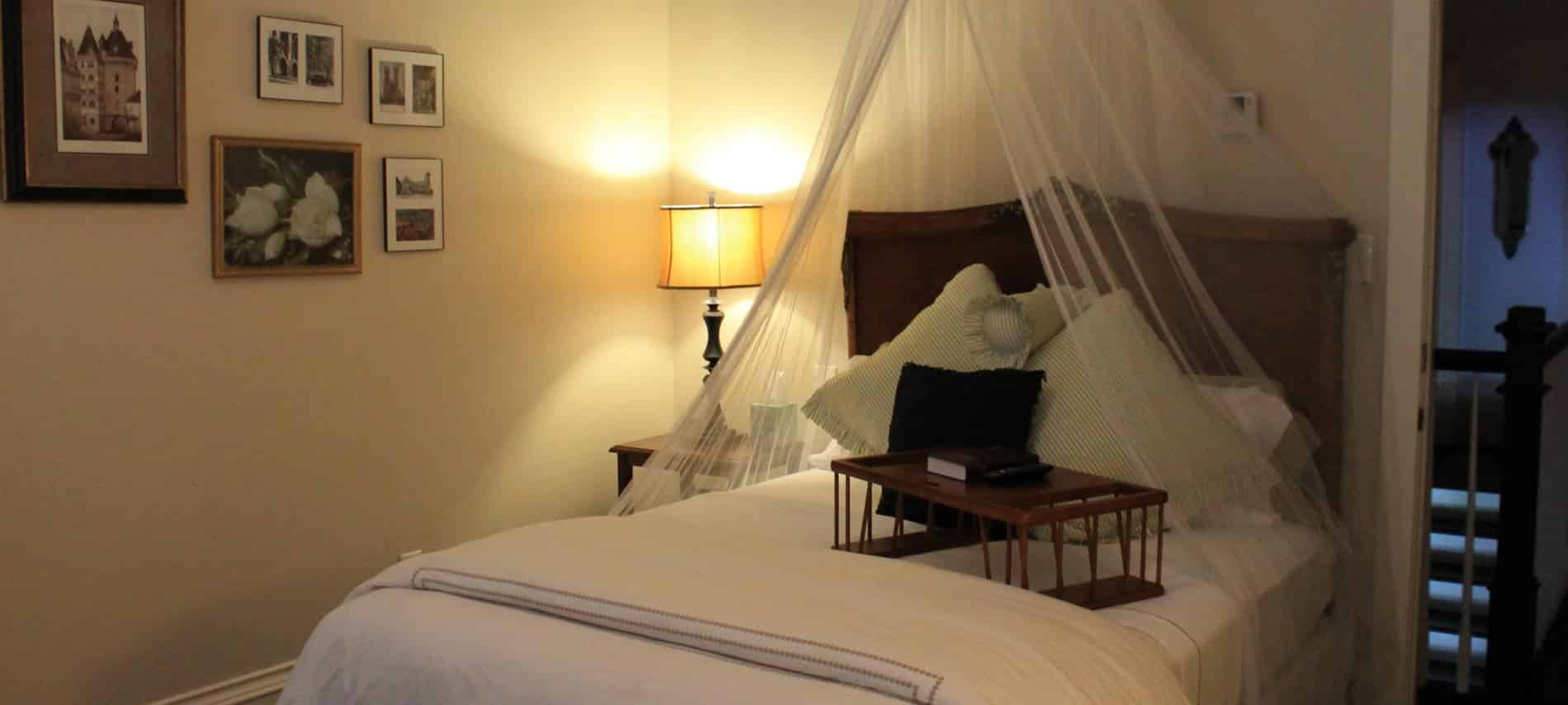 Charleston guest room, bed with tall wood headboard and sheer white canopy, nightstand with lamp, and white bedding