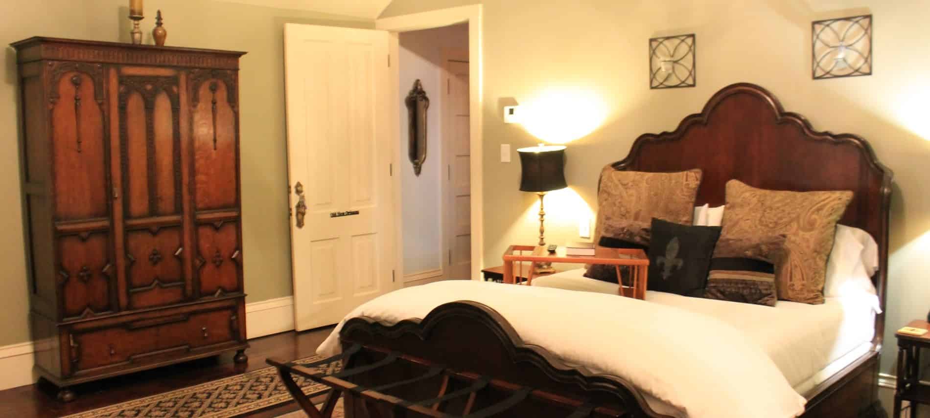 New Orleans guest room with light green walls, scalloped wood bed with white bedding, nightstand with lamps and tall armoire