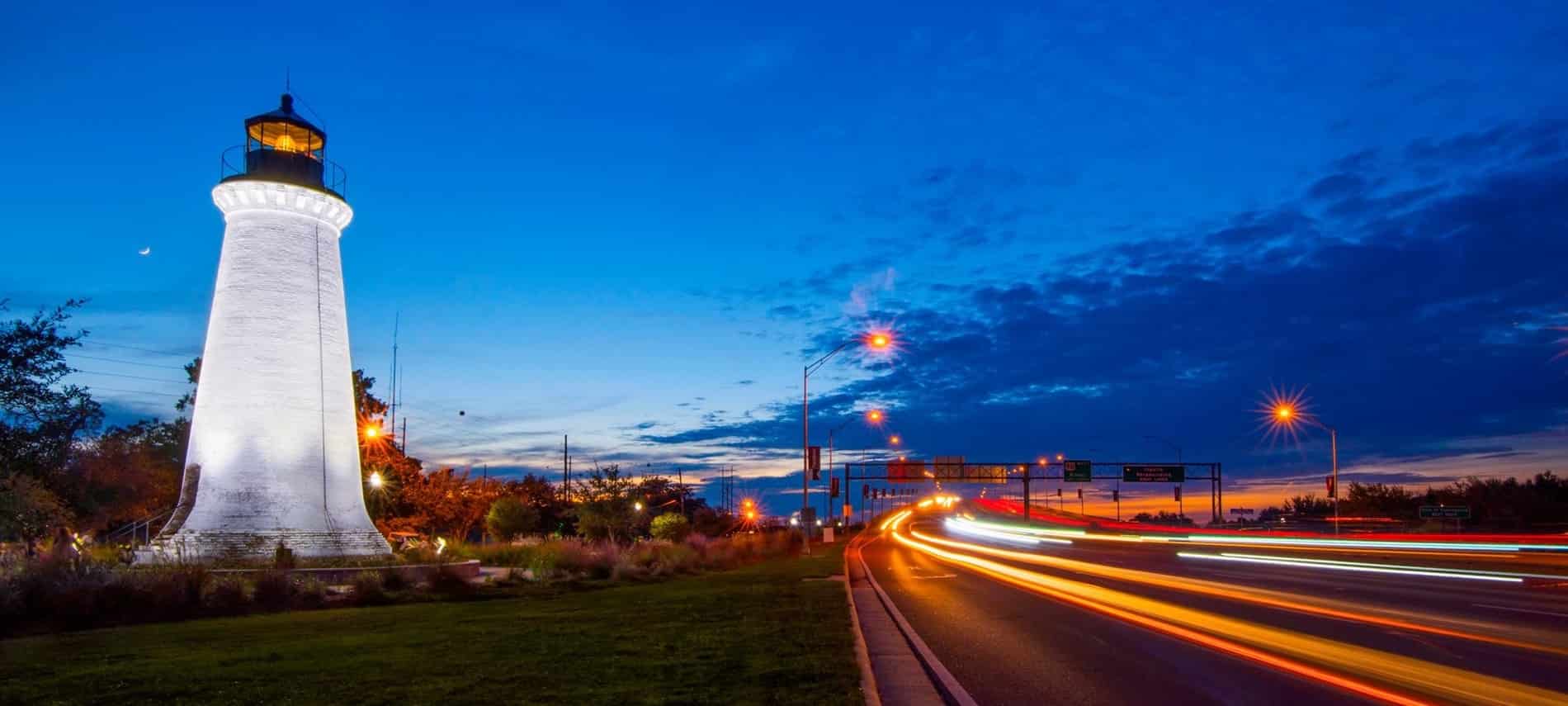 Exterior view of Round Island Lighthouse at dusk amidst deep blue skies with clouds and blurred city street and car lights