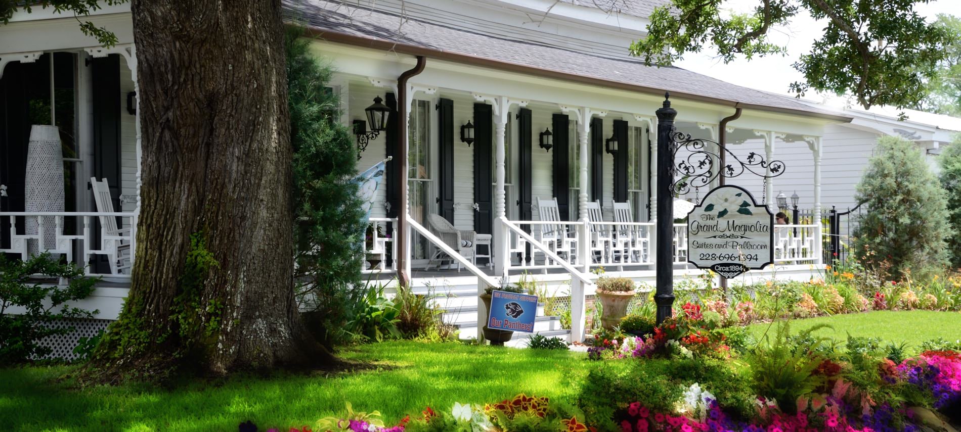 Exterior view, white sided home with covered porch, windows with black shutters amidst green grass, colorful flowers and mature tree