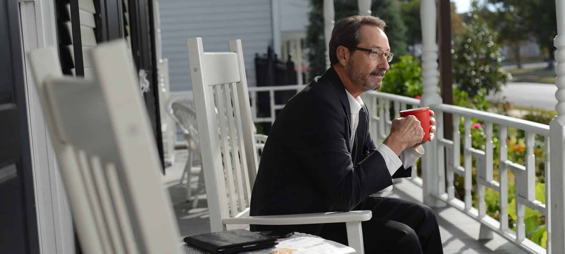 Man in black suit sitting in a white rocking chair on the front porch holding a red mug