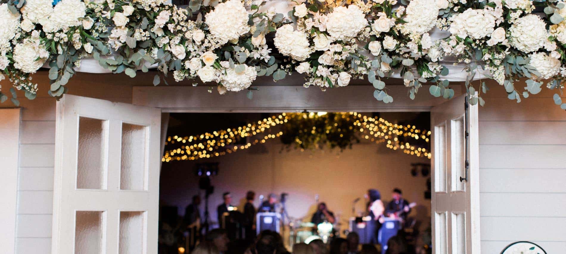 Double glass French doors swung open with large white flowers hanging overhead with view of wedding band and twinkle lights inside