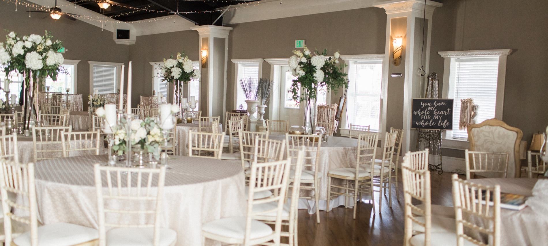 Beautiful ballroom with several windows decorated for a wedding with several ivory round tables and chairs topped with large white flowers s