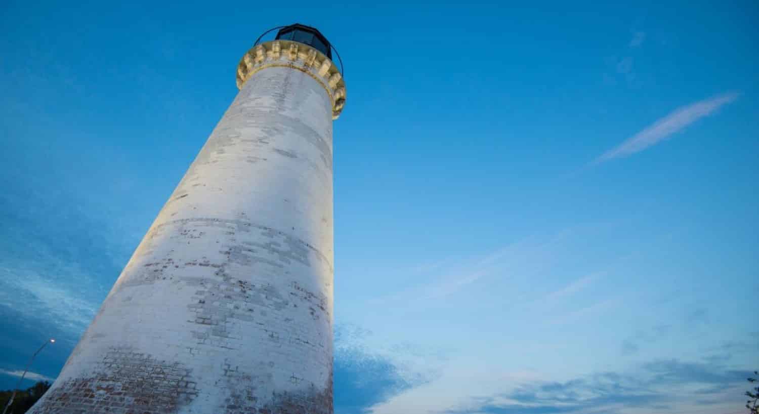 View of the white lighthouse from the base looking straight up amidst blue skies