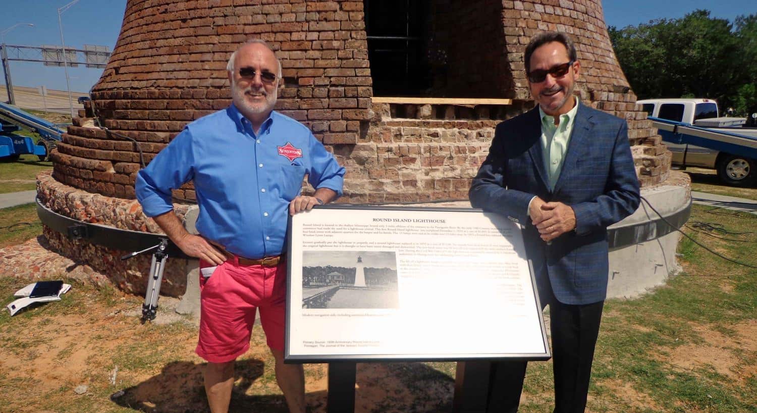 Two men standing on each side of the Round Island Lighthouse sign