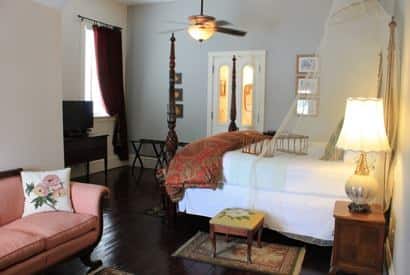 Savannah guest room with light walls, wood floor, dark four poster bed, ceiling fan, apricot sofa, and window