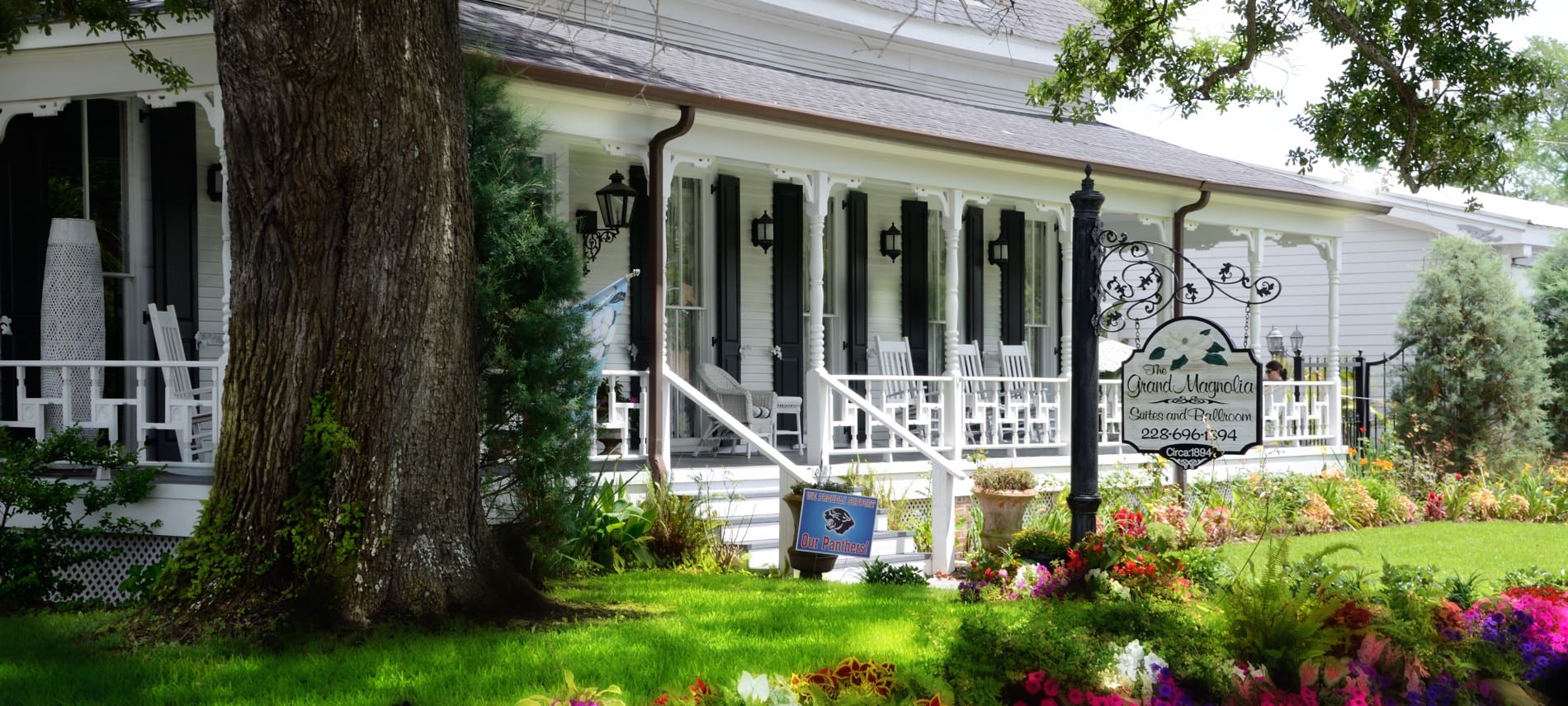 Exterior of Grand Magnolia with white siding, black shutters, covered porch, green lawn, mature trees and colorful flowers