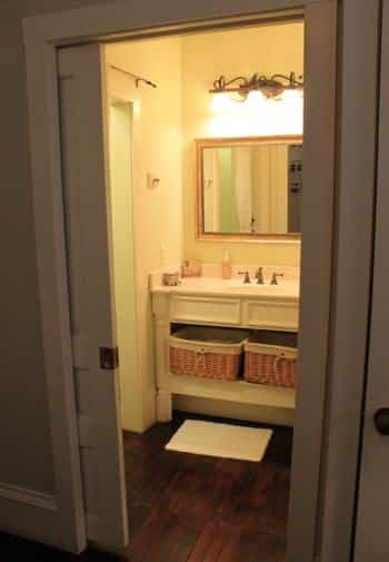 View of Charleston guest bath through a pocket door: white vanity with mirror, sconce lighting and shelf with baskets