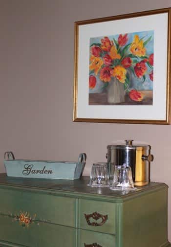 Garden guest room's green dresser topped with ice bucket and glass and picture of flowers in a gold frame overhead