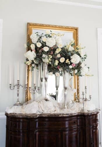 Dark wood console table topped with bunched white lace, two candelabras, two tall vases with flowers and gold rectangular mirror