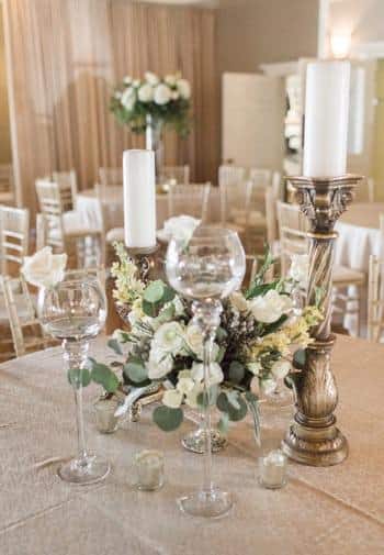 Elegant ivory chairs and round tables topped with ivory tablecloths, candleholders with white pillar candles and fresh white flowers