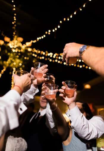 Several people from a bridal party raising glasses in a toast at night with twinkle lights overhead